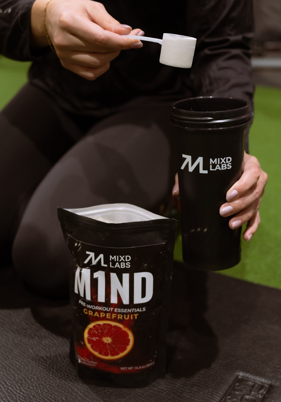 Person holding a black cup and package of M1ND by MIXD LABS on a green surface.