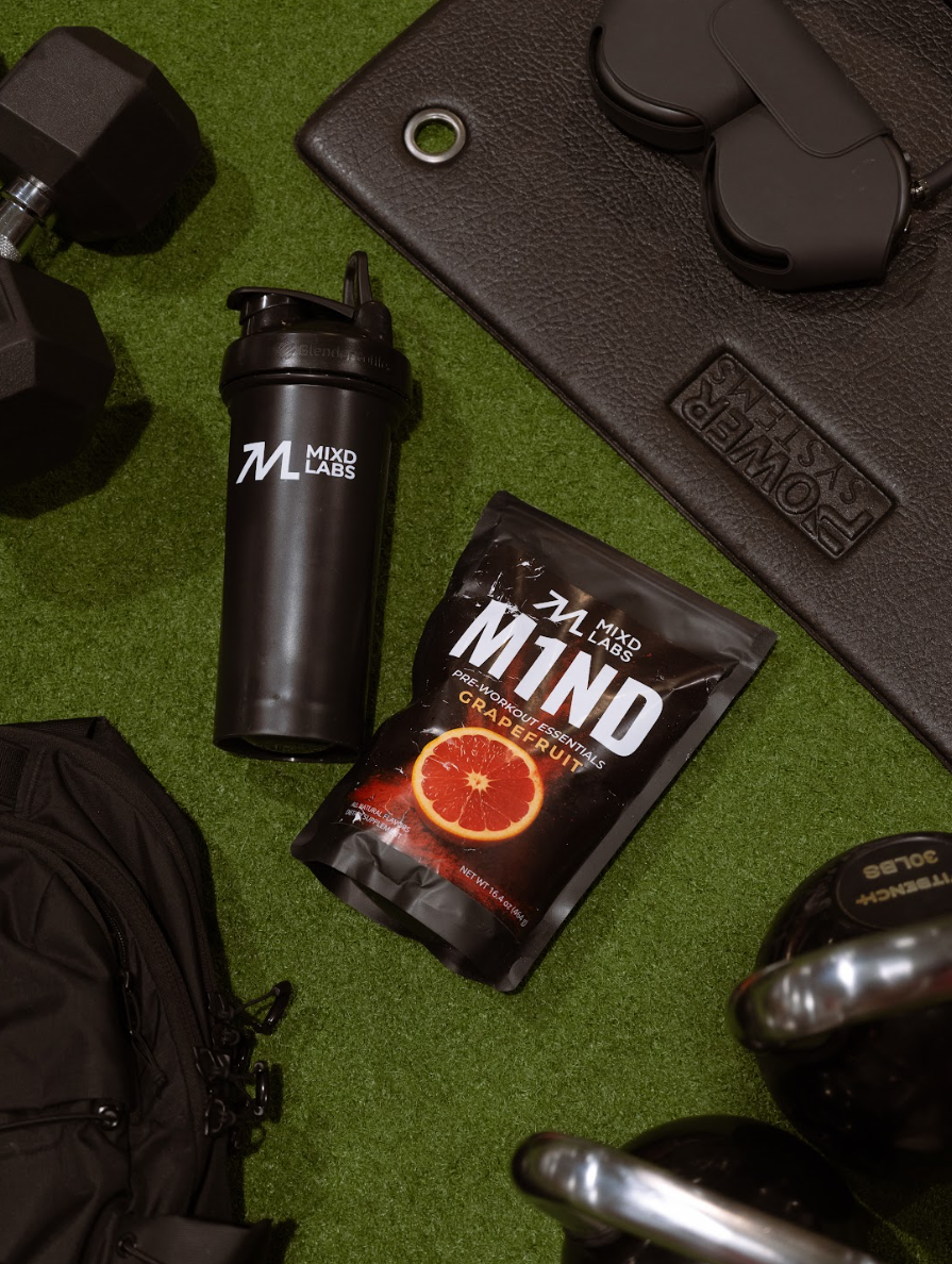 Black shaker bottle and supplement package on a green surface with gym equipment.